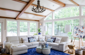 Modern cottage great room with exposed beams, classic blue rug and cream colored sectional surrounded by walls of windows.