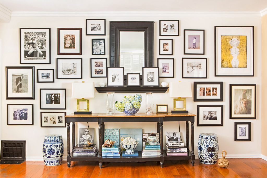 A dark console table against a large accent wall, covered with a large mirror, framed photographs and artwork. 