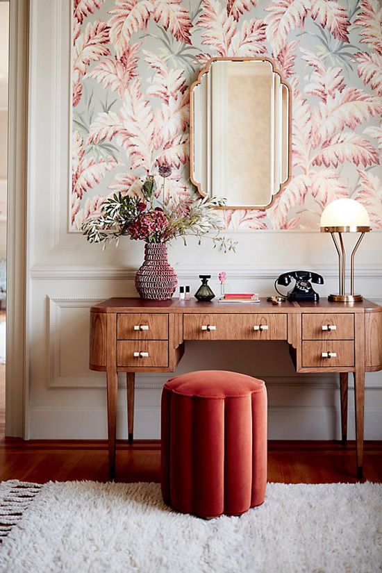 Velvet seat at a wooden vanity with topped with floral vase with an Art Deco styled mirror hanging on the wall. 