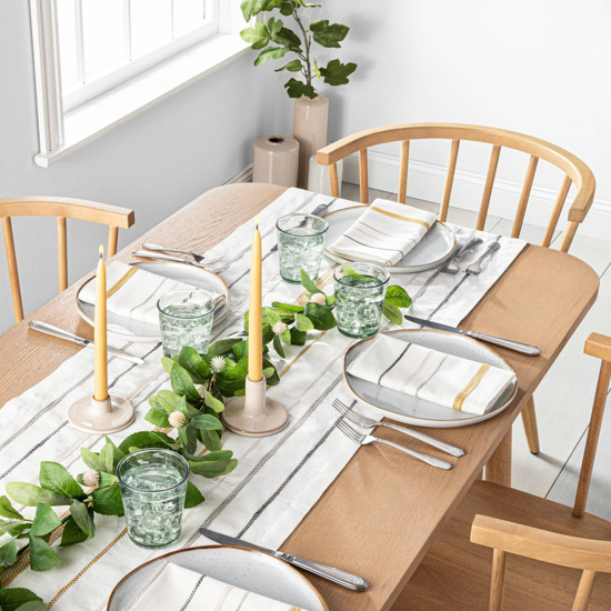 Light colored wooden table and chairs set for a meal with light colored table runner topped with yellow candles and fresh green garland. 