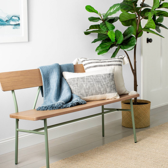 Green metal and wooden bench topped with a blue throw blanket and light colored pillows next to a fiddle leaf fig tree. 