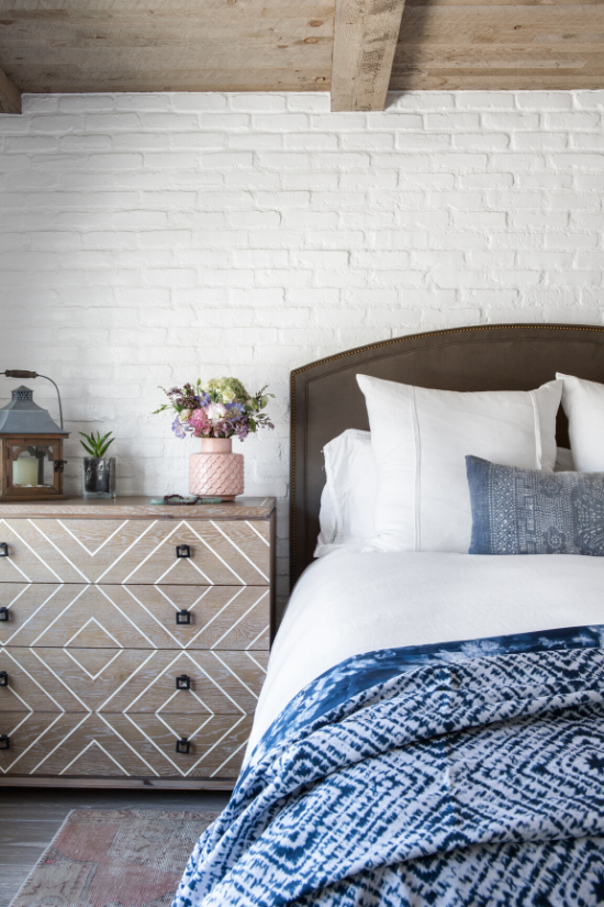 White brick wall and exposed wooden beam ceiling in a bedroom with a wooden dresser and a blue shibori comforter at the foot of the bed.