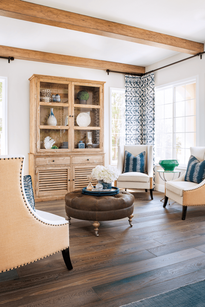 tall cottage sitting room with exposed beams, 4 slipper chairs a round tufted ottoman and a tall china cabinet in knotty pine