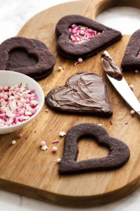 Wooden cutting board with heart shaped chocolate cookies, colorful Valentine's Day sprinkles and a knife covered in Nutella.