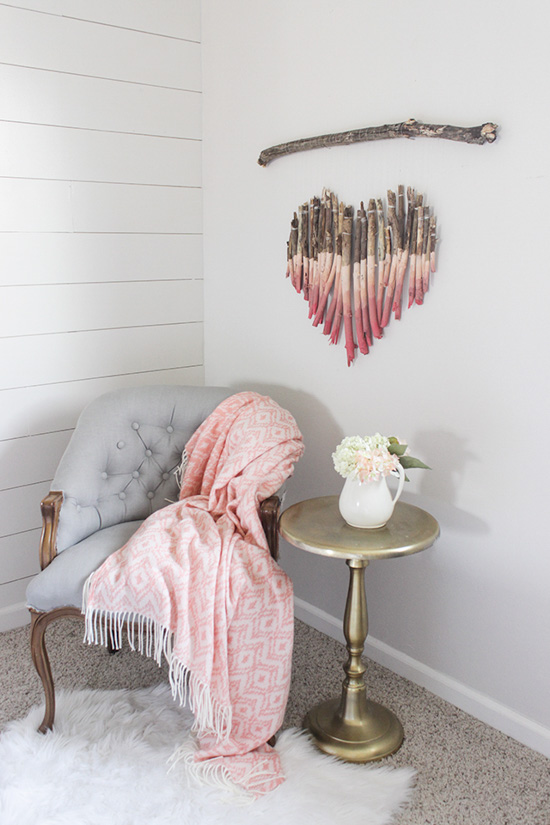 Tufted light gray chair, topped with a pink throw blanket and a handmade valentine's craft wall hanging in the shape of a heart made out of sticks. 