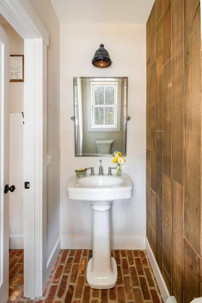 Small powder bath with white pedestal sink, brick flooring and wooden paneled accent wall. 
