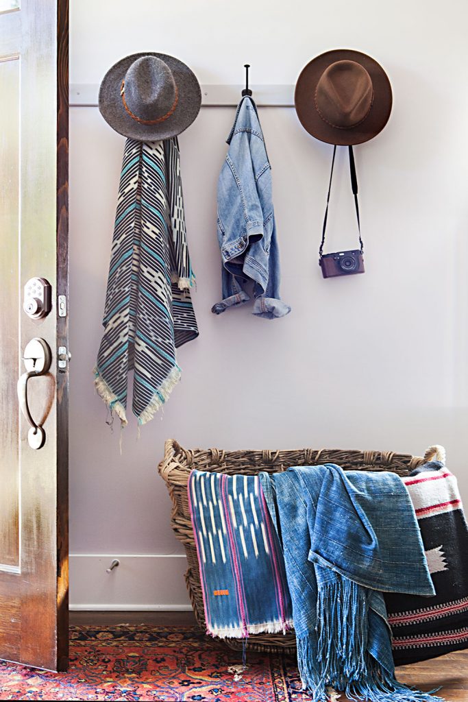 Declutter solution: stylish hooks and an oversized wide basket by the entryway of a Nashville cottage are filled with indigo textiles and stylish denim jackets, wide brimmed hats and a mexican poncho 