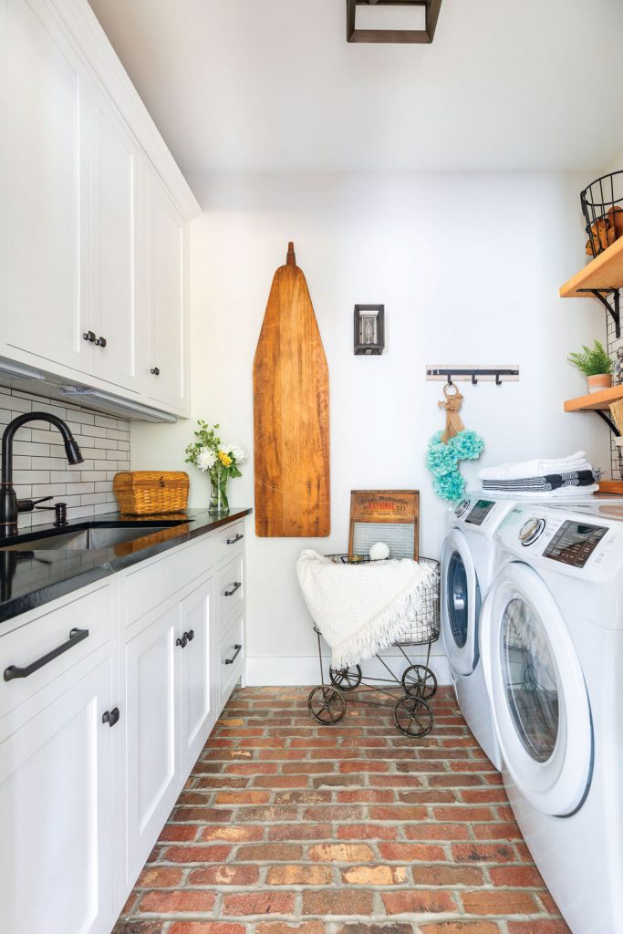 Laundry room with brick flooring, white washer and dryer, open shelving and wooden ironing board. 