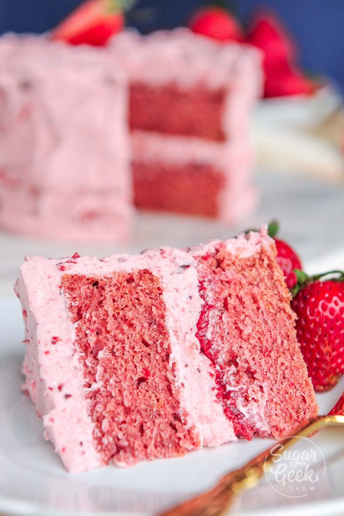 Slice of strawberry cake plated with fresh strawberries and a gold fork.