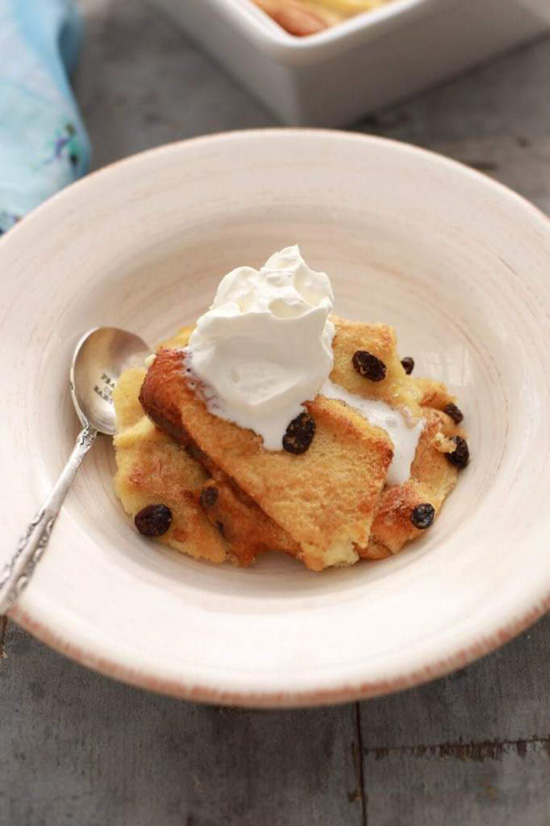 Cream colored dish with a silver spoon and a serving of bread pudding topped with whipped cream. 