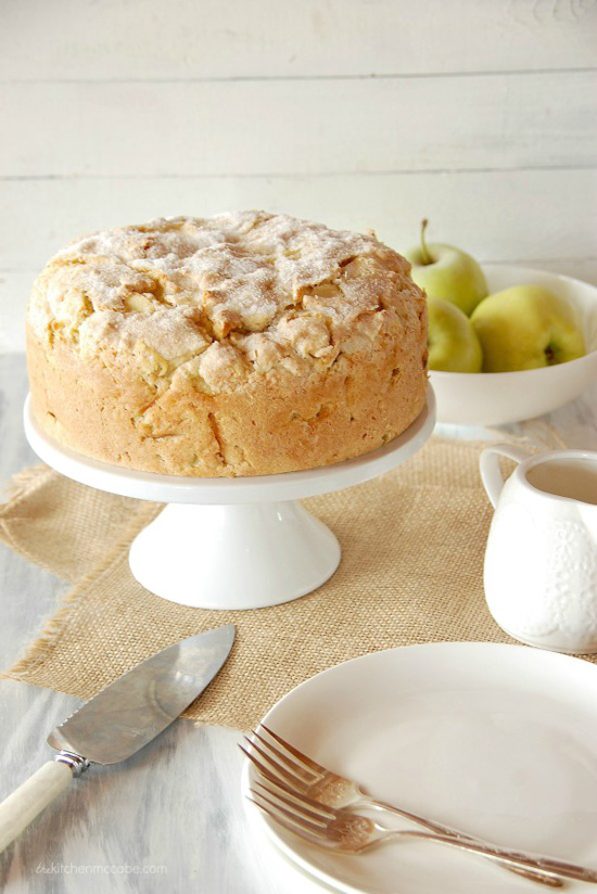 Irish apple cake displayed on a white cake stand laid on linen table cloth and bowl filled with green apples. 