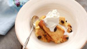 Cream colored dish with a silver spoon and a serving of bread pudding topped with whipped cream.