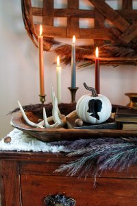 Painted silhouette pumpkin in wood dough bowl with books, candles, and shed antlers