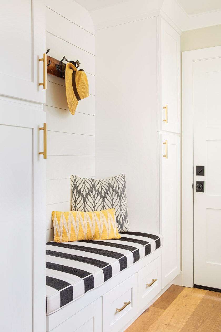 white mudroom/storage area with black and white striped bench cover and yellow pillow and straw hat hanging above 