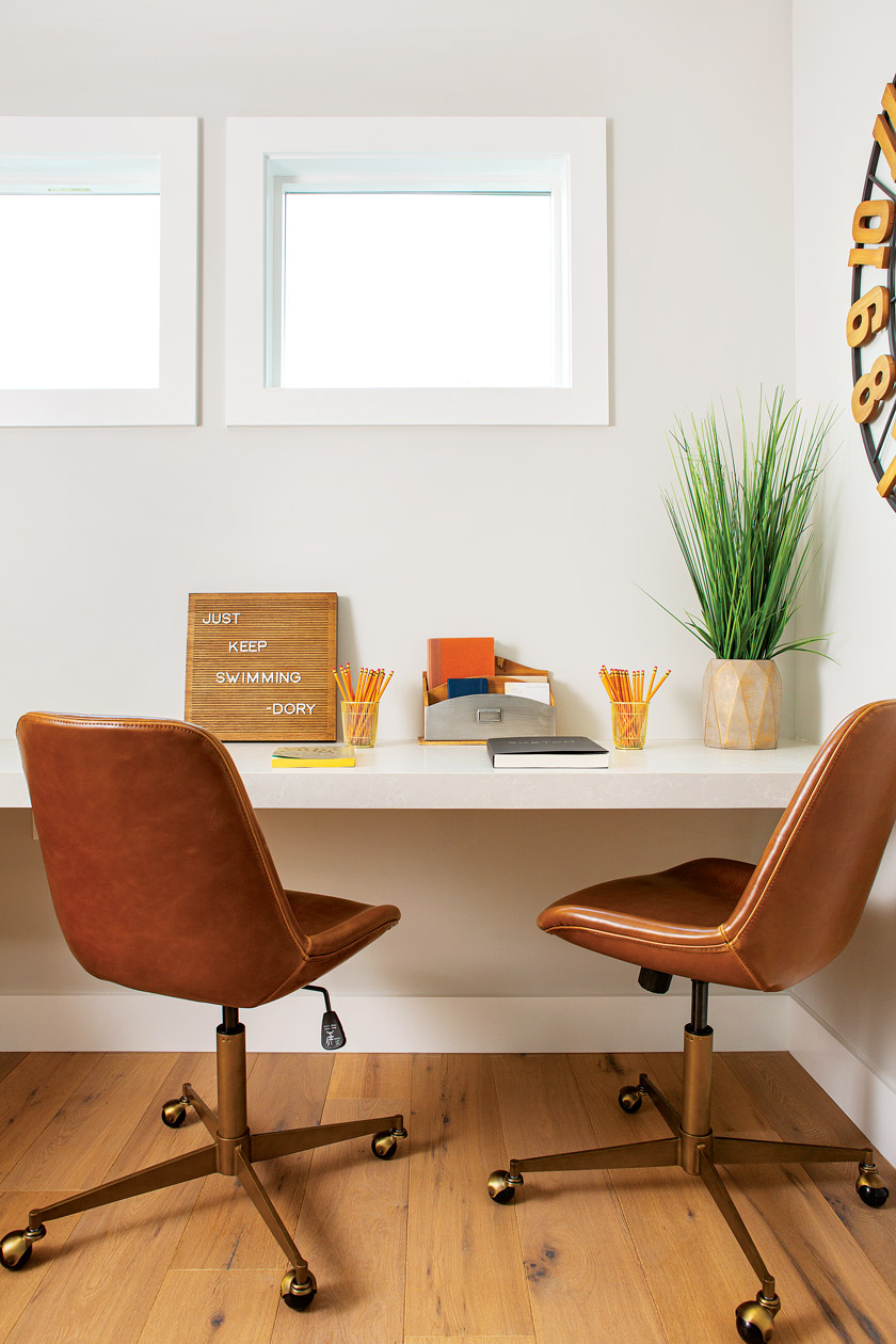 desk area in a coastal farmhouse with two brown leather swivel chairs beneath two smaller windows