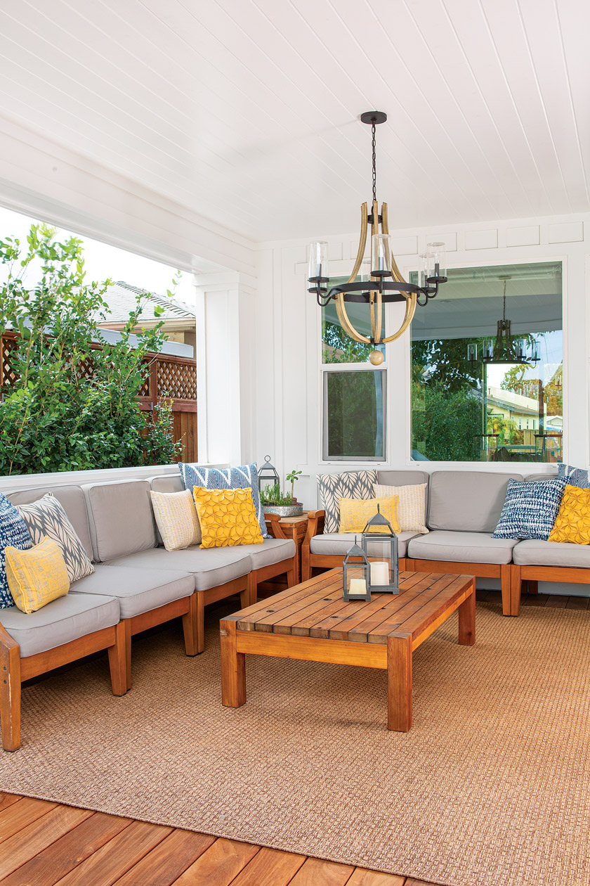 open patio with large sectional seating with yellow and coastal blue pillows and teak table with large chandelier above 