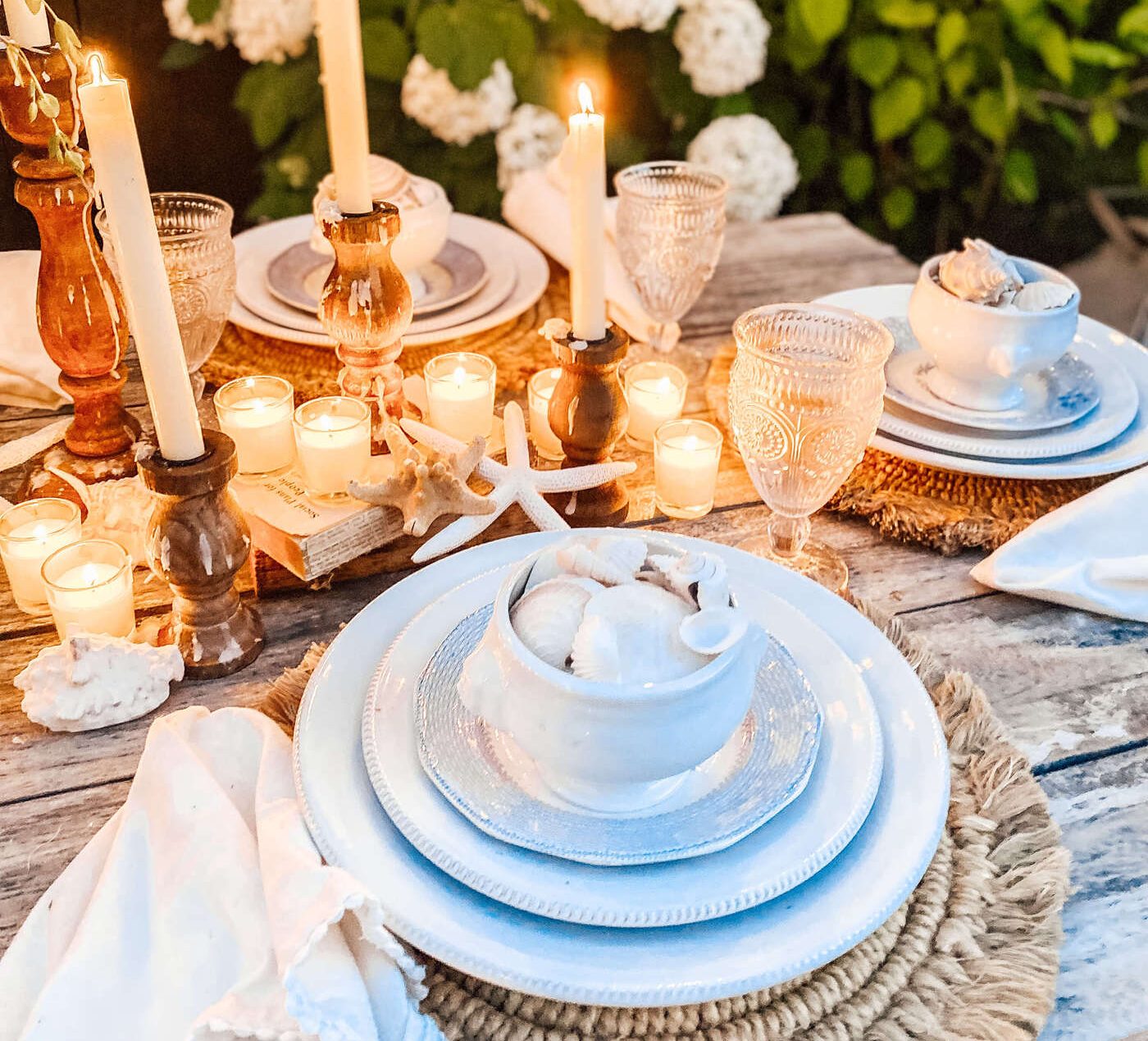 Beach table setting with white ceramic dishes, tea candles, white shells, and rattan chargers