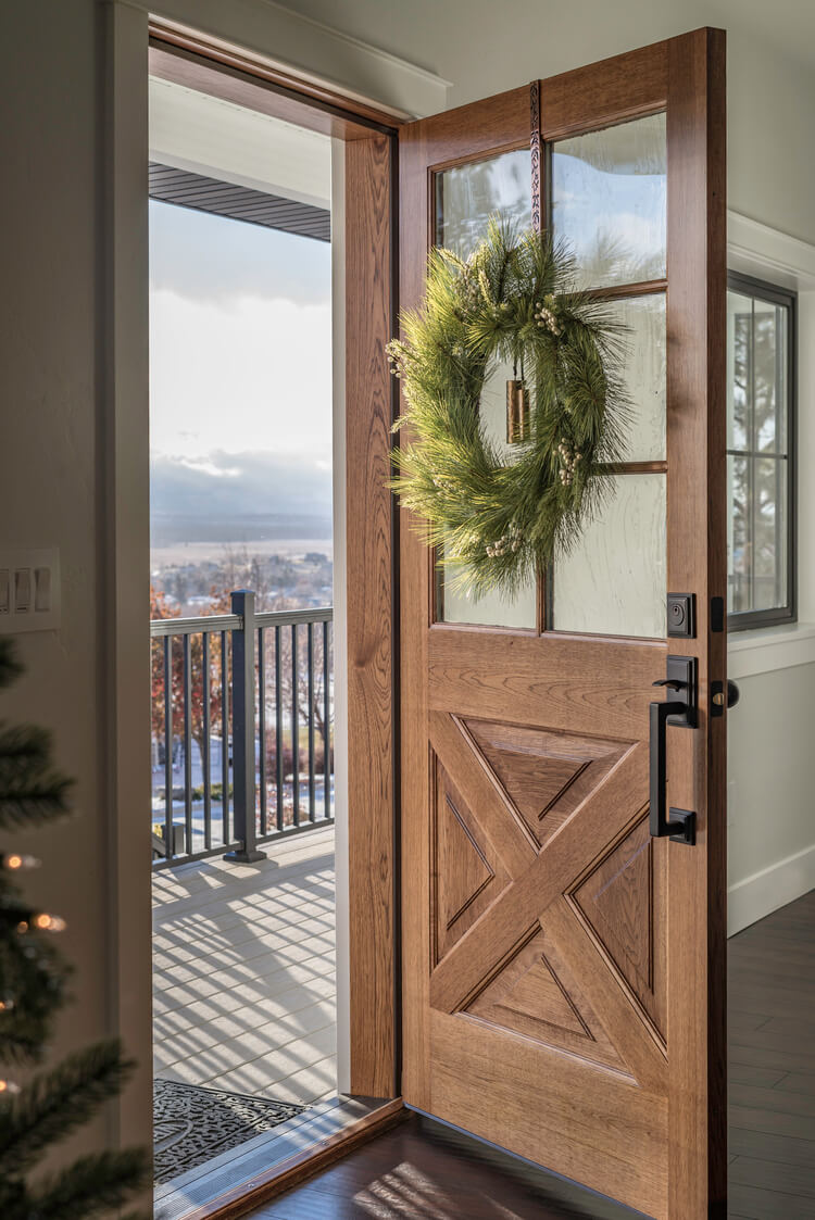 A wreath hangs on an open wood door that has black hardware and glass panels