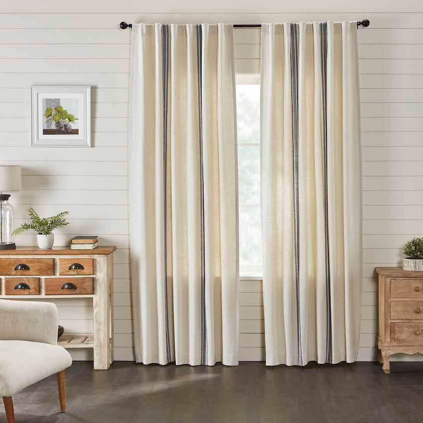 Between two natural wood side tables is a window with cream curtains and thin blue stripes.