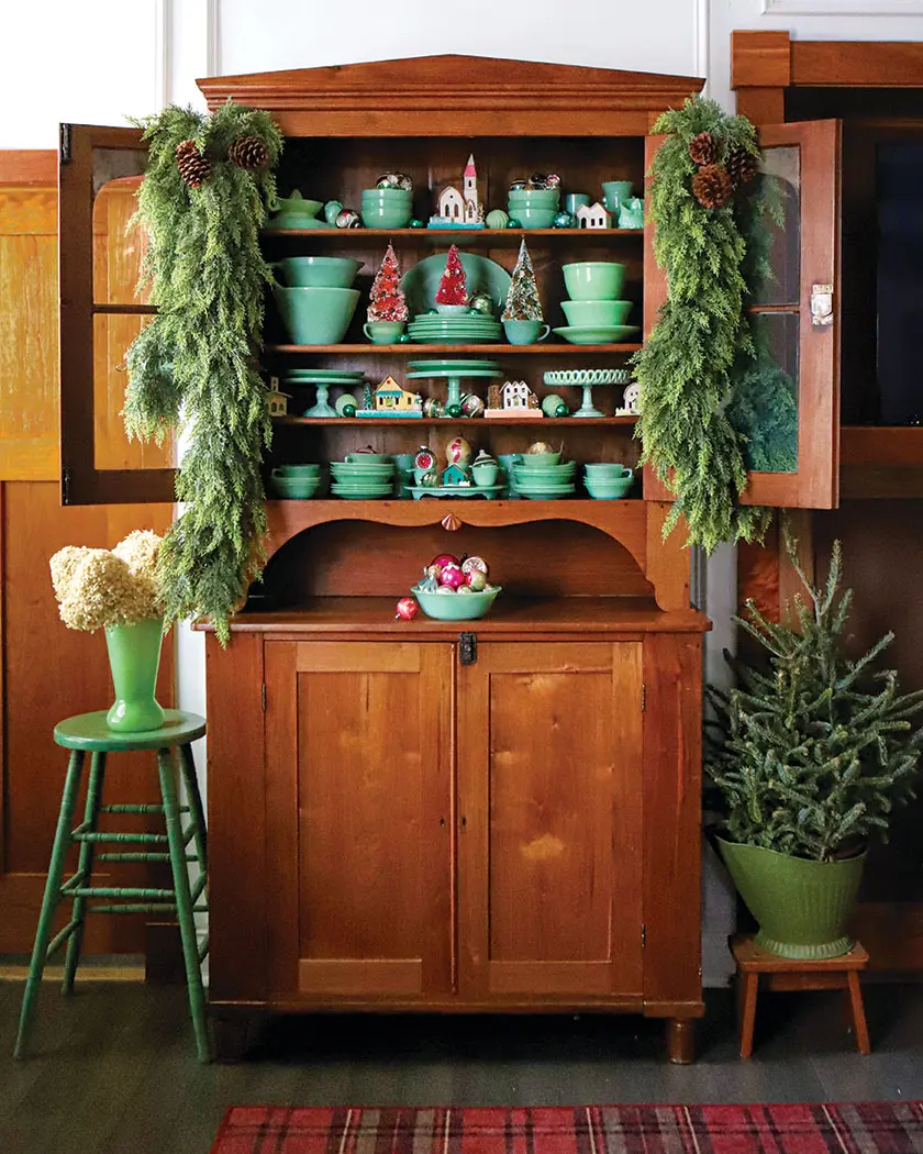 fresh greenery on hutch containing jadeite collection putz houses and shiny brite ornaments