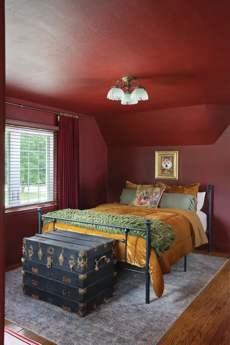 deep maroon painted walls and iron bedframe with saffron and green bedding and antique trunk at the foot of the bed for storage